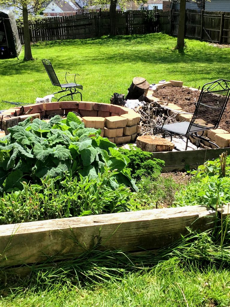 Aromatic herbs in a raised bed, adjacent to a large brick firepit and a new raised bed vegetable garden.